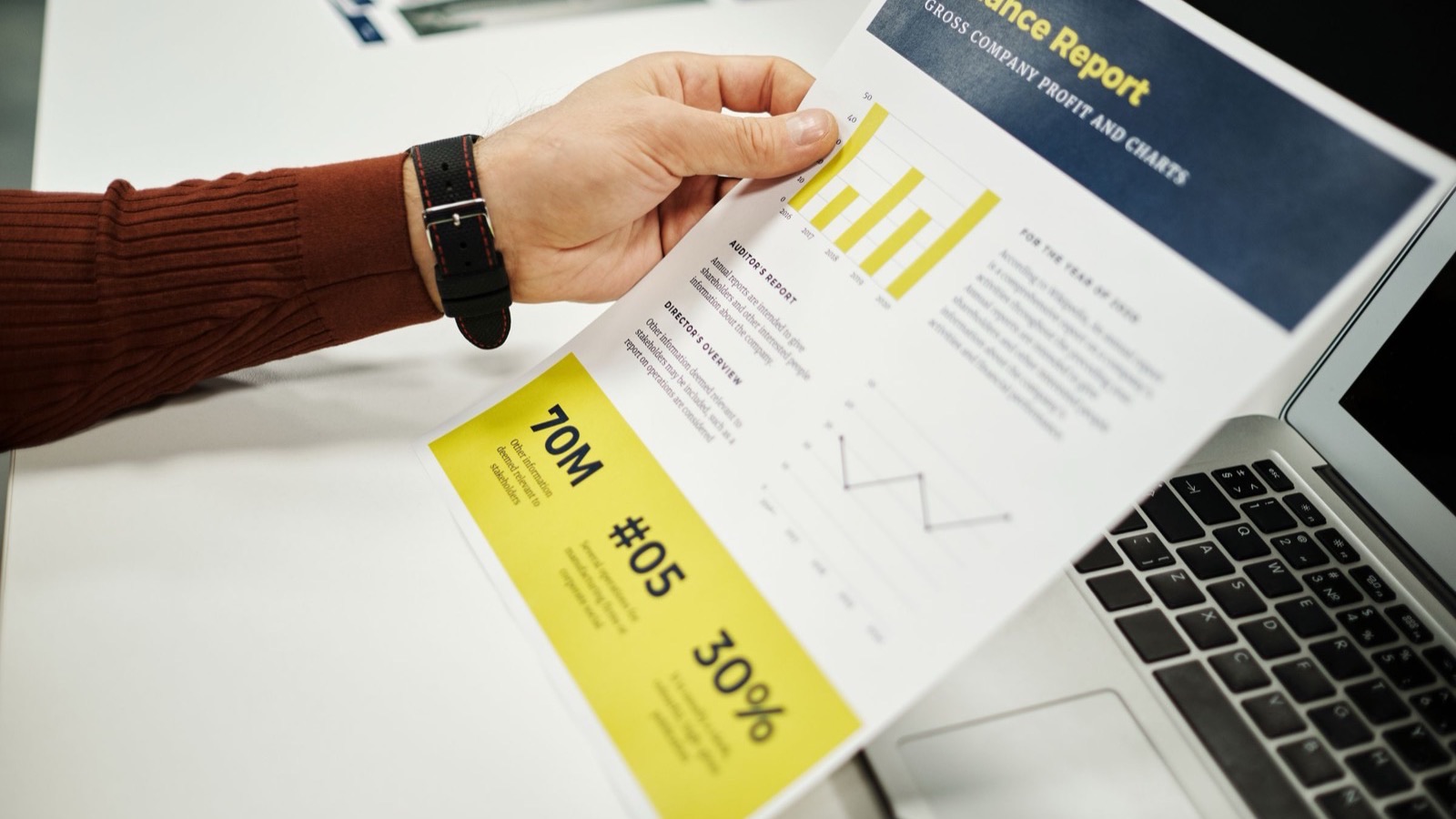 A hand holding a printed performance report with bar charts and profit figures, beside a laptop on a desk