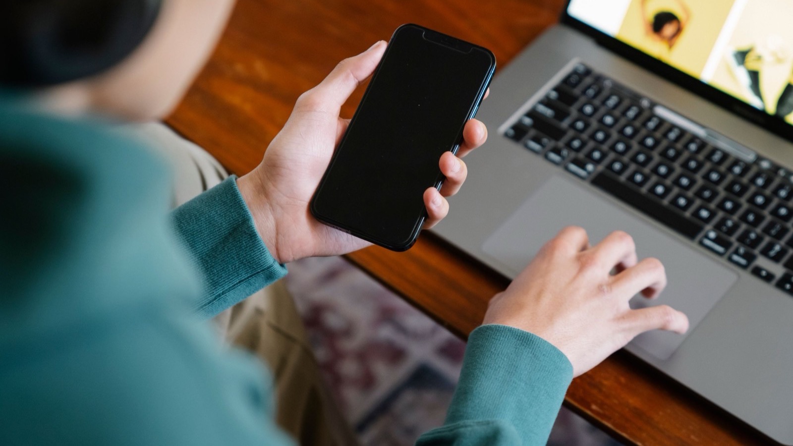 Someone holding a smartphone in one hand while typing on a laptop at a wooden desk