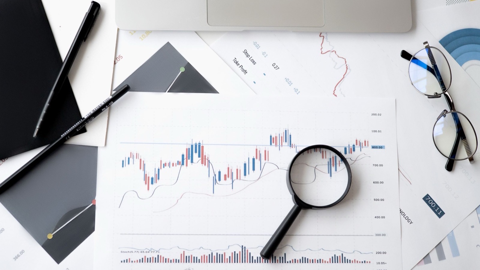 An overhead view of printed financial charts with a magnifying glass resting on them, beside pens, reading glasses, and a laptop